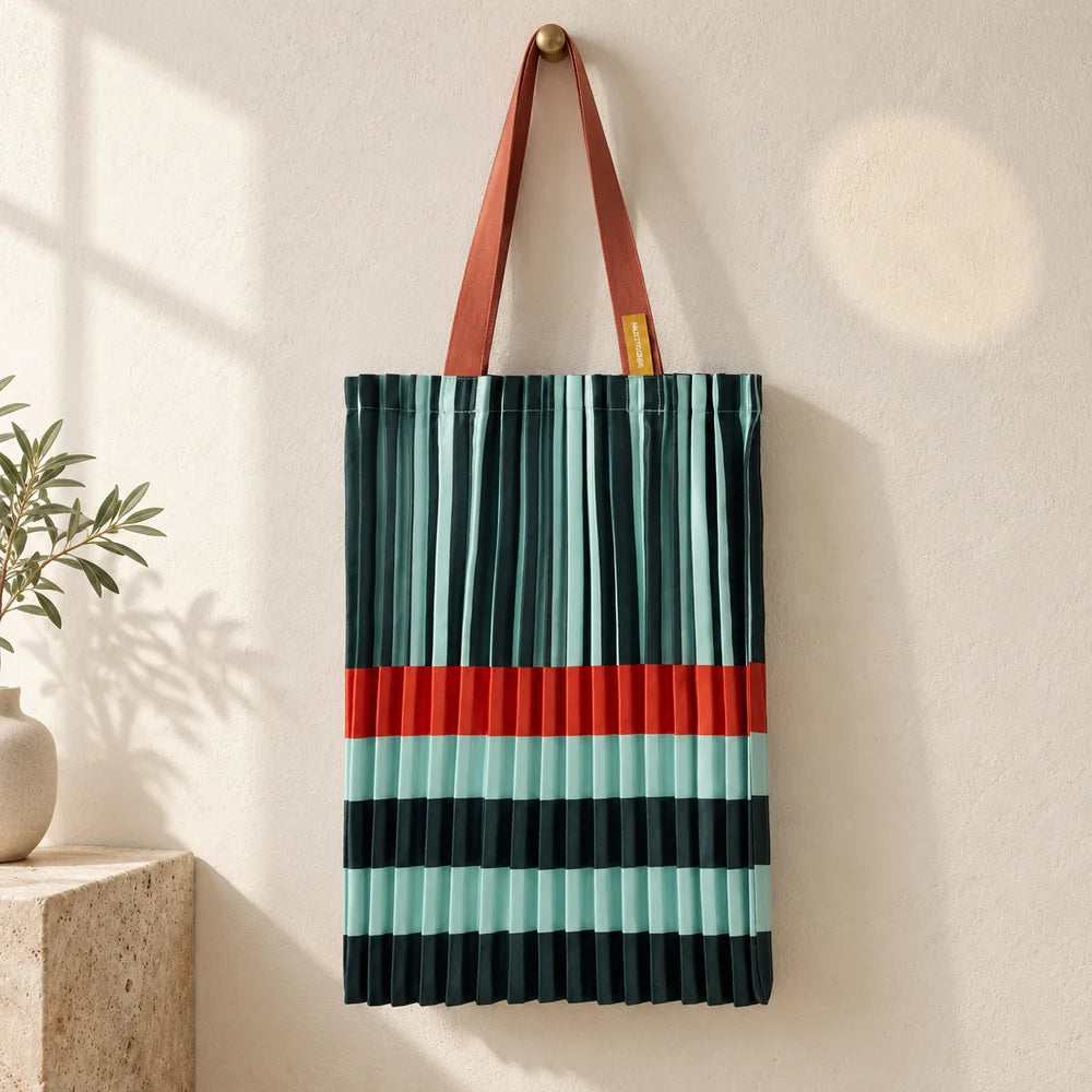 Tote plissettata verde e acqua con fascia arancio e manici ruggine, appesa su una parete avorio.