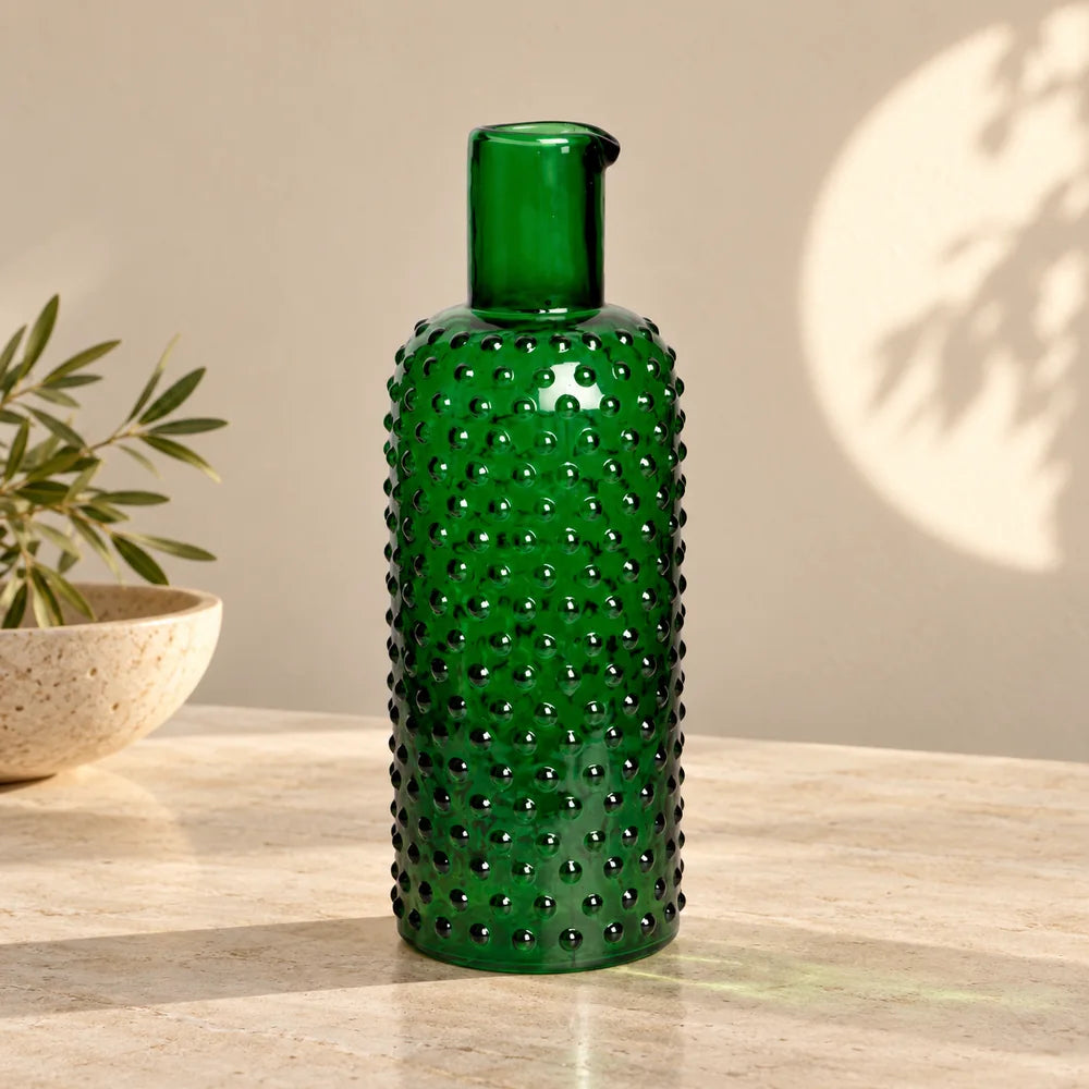 Caraffa verde scuro in vetro a rilievi su pietra chiara, senza manico.