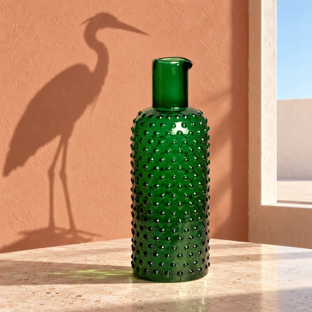 Caraffa verde su una tavola chiara, con l’ombra inattesa di un airone sulla parete.