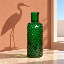 Caraffa verde su una tavola chiara, con l’ombra inattesa di un airone sulla parete.