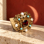 Anello d’Este su un piano avorio, con pietra rossa centrale, quattro perle bianche e piccoli dettagli turchesi.