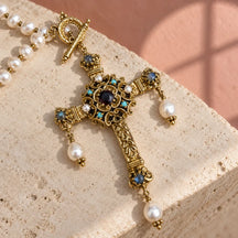 Croce Castel in ottone dorato con pietre blu, cristallo scuro e tre perle pendenti, su pietra avorio.