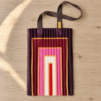 Tote plissettata amaranto con rettangoli rosa e crema, distesa su legno chiaro.