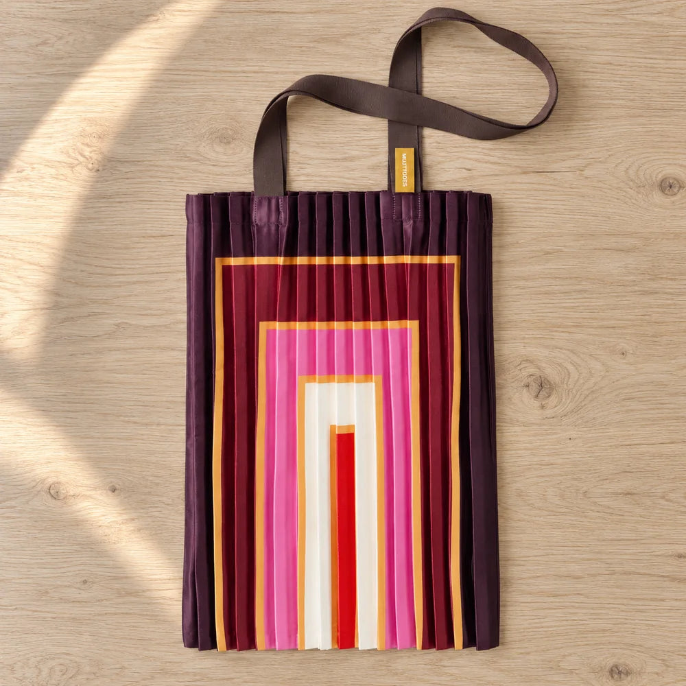 Tote plissettata amaranto con rettangoli rosa e crema, distesa su legno chiaro.