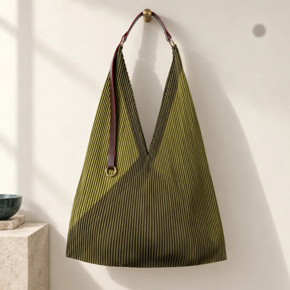 Borsa verde oliva plissettata con manico in pelle e anello dorato, appesa su parete avorio.