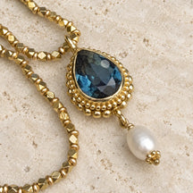 Pendente Bisanzio con cristallo London Blue a goccia e montatura dorata su travertino chiaro.