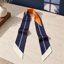 Twilly blu scuro e arancio in seta appoggiato su pietra avorio, con entrambe le facce e le punte bianche visibili.